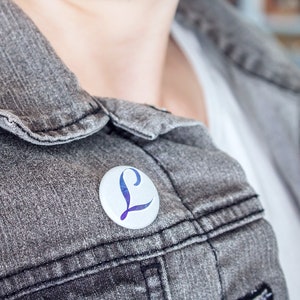 Könnte beinhalten: Eine graue Jeansjacke mit einem weißen Knopf und einer blauen und violetten Anstecknadel mit dem Buchstaben L.