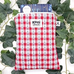 Könnte beinhalten: Eine rot-weiß karierte Bucheinbandhülle mit weißem Reißverschluss. Die Hülle hat ein Etikett mit der Aufschrift "kuni + plum + handmade". Die Hülle enthält ein Buch mit blauem Einband und dem Titel "Joyous" von Neil Gaiman.