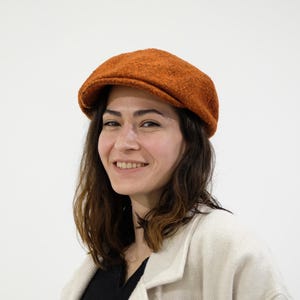 May include: A burnt orange newsboy cap. The cap has a rounded crown and a small, stiff brim. The fabric appears to be a textured wool or tweed. The person wearing the cap is smiling and wearing a cream-coloured coat.