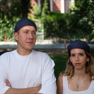 May include: Two individuals wearing matching dark blue beanies. One is in a white t-shirt, arms crossed. The other wears a white vest top and silver necklace. They stand before a green backdrop of trees and foliage.