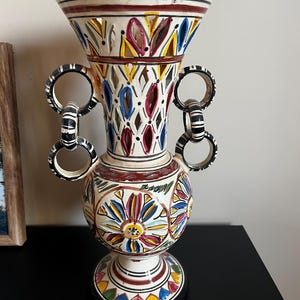 Può includere: Un vaso in ceramica con base color crema e motivi floreali dipinti a mano in rosso, blu e giallo. La parte superiore presenta un design traforato, mentre i lati includono due manici con anelli pendenti decorati in bianco e nero.