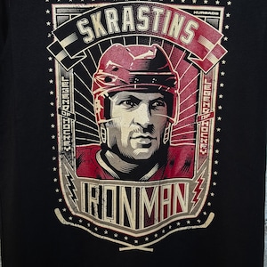 Pu&ograve; includere: Maglietta nera con una stampa grafica di un giocatore di hockey con un casco rosso. La grafica include il testo "SKRASTINS", "LEGEND OF HOCKEY", "IRONMAN" e "KURBADS".