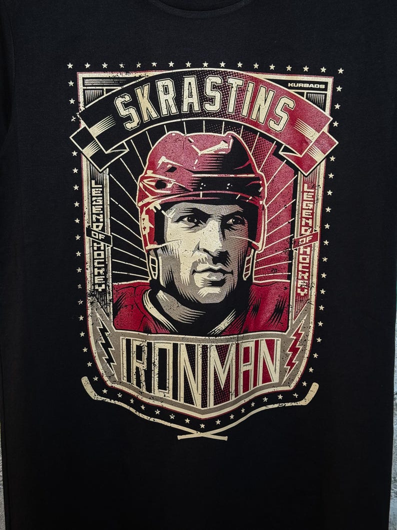 Pu&ograve; includere: Maglietta nera con una stampa grafica di un giocatore di hockey con un casco rosso. La grafica include il testo "SKRASTINS", "LEGEND OF HOCKEY", "IRONMAN" e "KURBADS".