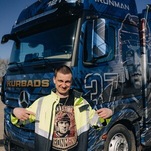 Pu&ograve; includere: Un uomo indossa una maglietta nera con una grafica di un giocatore di hockey e il testo "RASTINS IRONMAN" e si trova davanti a un camion semi-rimorchio blu con la scritta "IRONMAN" sul lato. Il camion &egrave; decorato con un motivo mimetico blu, bianco e nero.