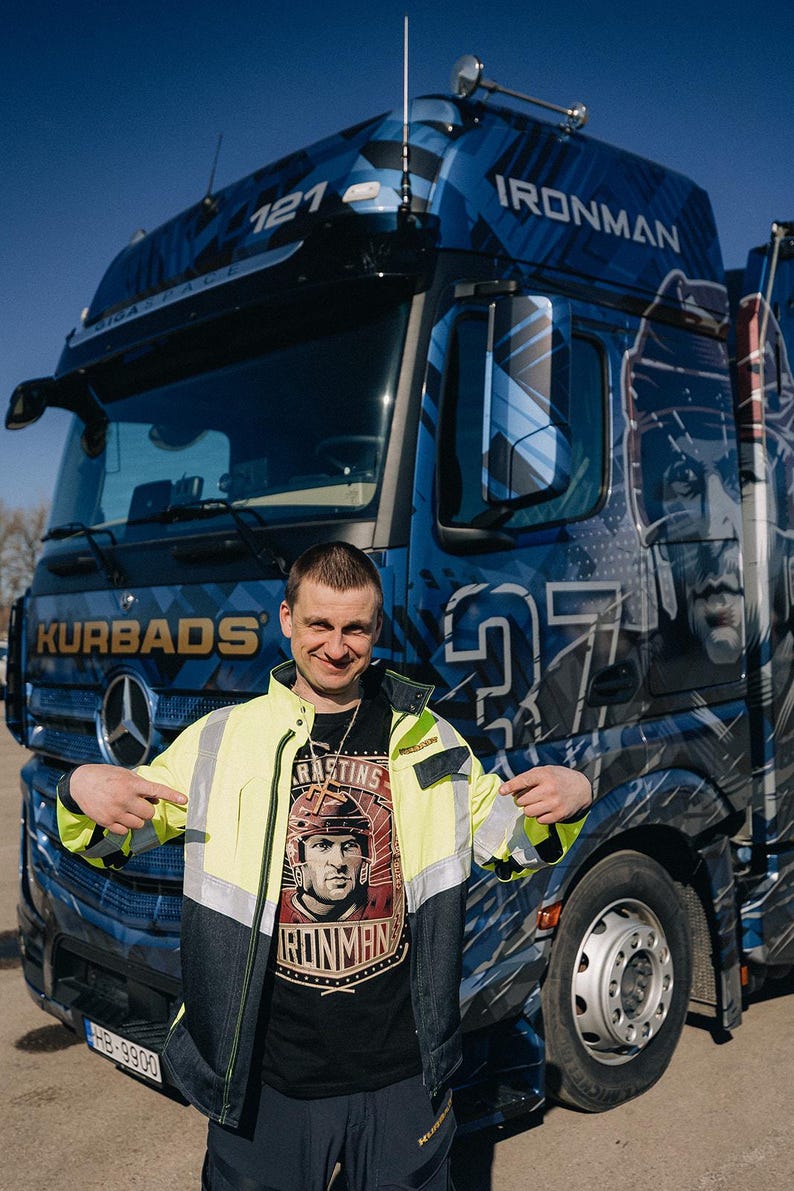 Pu&ograve; includere: Un uomo indossa una maglietta nera con una grafica di un giocatore di hockey e il testo "RASTINS IRONMAN" e si trova davanti a un camion semi-rimorchio blu con la scritta "IRONMAN" sul lato. Il camion &egrave; decorato con un motivo mimetico blu, bianco e nero.
