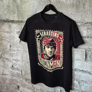 Pu&ograve; includere: Maglietta nera con una stampa grafica di un giocatore di hockey che indossa un casco e una maglia rossi. La grafica include il testo "SKRASTINS" e "IRONMAN".
