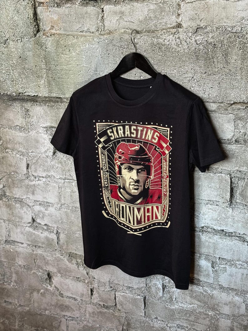Pu&ograve; includere: Maglietta nera con una stampa grafica di un giocatore di hockey che indossa un casco e una maglia rossi. La grafica include il testo "SKRASTINS" e "IRONMAN".