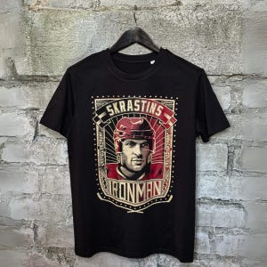 Peut inclure: T-shirt noir avec un graphique d'un joueur de hockey portant un casque et un chandail rouges. Le graphique comprend le texte "Skrastins" et "Ironman".