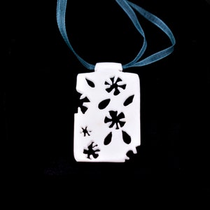 May include: White ceramic pendant necklace with a floral design. The pendant is shaped like a rectangle with cutouts of flowers and leaves. The necklace is on a teal ribbon.
