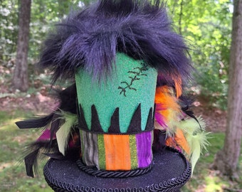May include: A black top hat with a green, orange, and purple striped band. The hat has a black feather trim and a black fuzzy top. The hat is sitting on a white stand. "Brown Girls Designs" is written on the stand.