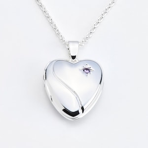 May include: A silver heart-shaped locket necklace with a fine chain. The locket has a swirling design and a small purple gemstone. The necklace is shown against a white backdrop.