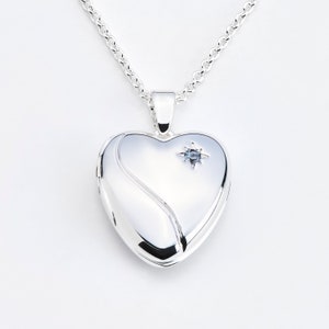May include: A silver heart-shaped locket necklace with a small blue gemstone starburst accent. The locket hangs from a delicate silver chain. The locket has a smooth, polished surface with a curved design element on the front.