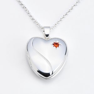 Könnte beinhalten: Silberner herzförmiger Anhänger mit Kette. Das Medaillon hat ein dekoratives Wirbeldesign und einen kleinen orangefarbenen Edelstein. Kette und Anhänger bestehen aus glänzendem, reflektierendem Metall.