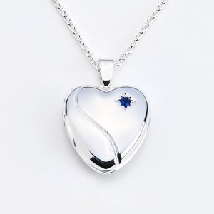 May include: A silver heart-shaped locket pendant on a chain. The locket has a polished finish with a curved design on the front and a small blue gemstone. The chain is also silver.