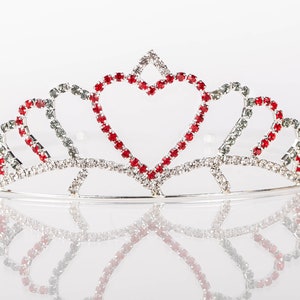 以下が含まれることがあります： ハート型のデザインが施されたシルバーのティアラ。赤いラインストーンとクリアなラインストーンが特徴で、中央のハートは赤で縁取られています。白い表面にティアラが映っています。
