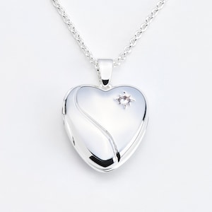 May include: A silver heart-shaped locket pendant on a chain. The locket has a curved design on the front and a small, star-shaped gemstone. The chain is a delicate, linked style.
