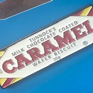 May include: A red, white, and yellow enamel pin shaped like a Tunnock's Caramel wafer biscuit. The pin features the text "Tunnock's Coated Chocolate Caramel Wafer Biscuit" and the Tunnock's logo.