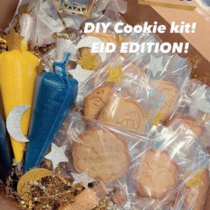 May include: A DIY cookie kit for Eid, featuring a box filled with cookie dough, icing bags, sprinkles, and gold and silver stars. The box also includes a small glass jar filled with gold beads. The text on the box reads "DIY Cookie Kit! Eid Edition!"