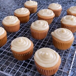 May include: A close-up of a wire rack with a dozen cupcakes frosted with a swirl of white frosting. The cupcakes are arranged in rows on the rack.