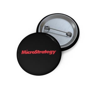 Könnte beinhalten: Zwei runde schwarze Ansteckbuttons. Ein Button zeigt den roten Text "MicroStrategy". Der andere Button ist mit der silbernen Rückseite nach unten.