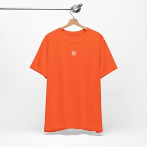 May include: Orange short-sleeved t-shirt with a small white circle containing the letter "B" on the front.