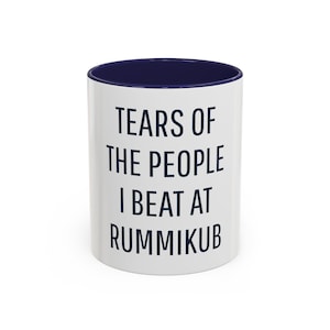 May include: White ceramic coffee mug with a navy blue rim and text that reads "Tears of the people I beat at Rummikub".