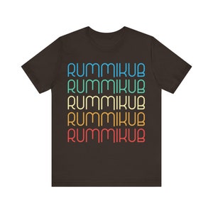 Puede incluir: Una camiseta marrón oscuro con la palabra "Rummikub" repetida cinco veces en un arcoíris de colores. El texto está apilado verticalmente, con cada palabra en un color diferente.