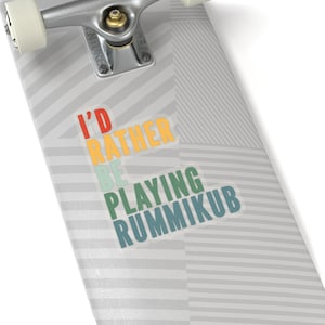 May include: A skateboard with a sticker that reads "I'd rather be playing Rummikub". The sticker has a retro color scheme with red, orange, yellow, green, and blue.