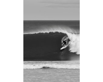 May include: Black and white photograph of a surfer riding a large wave. The surfer is silhouetted against the wave's curl, with the ocean and sky in the background. The image is printed on a poster and held by two black binder clips.