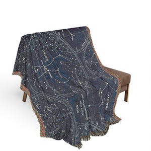 May include: A dark blue woven throw blanket featuring a detailed celestial map with white star constellations, lines, and text including Orion, Cassiopeia, and Milky Way. The blanket has a textured finish and frayed edges along the bottom border.