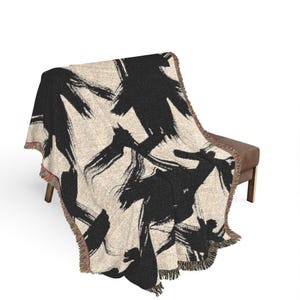 May include: A woven throw blanket featuring a cream-colored base with bold, black abstract brushstroke patterns. The blanket has a textured appearance and is finished with frayed, multi-colored fringe edges along the sides and bottom.