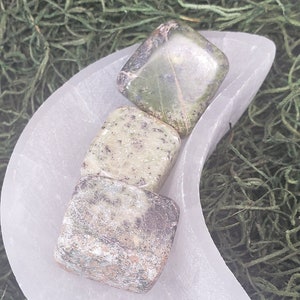 May include: Three square-shaped stones in shades of green, gray, and brown rest on a white, crescent-shaped dish. The dish sits on a bed of green, textured material, creating a natural, earthy aesthetic. The stones appear to be crystals or minerals.