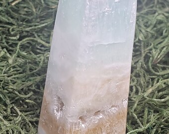 May include: A layered crystal tower with a polished, rectangular shape. The crystal features bands of white, light blue, and tan colors. The tower is set against a backdrop of green moss.