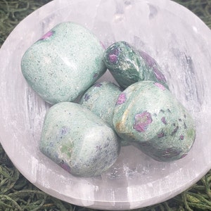 May include: A collection of tumbled ruby in zoisite stones, displaying a mix of green and pink hues. The stones are arranged in a white, shallow bowl, set against a backdrop of green moss.