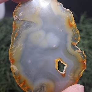 May include: A polished agate stone slice, irregularly shaped. It has a central grey area with white and yellow banding, and a small, diamond-shaped hole. The outer edge features orange and yellow hues.