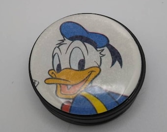 Puede incluir: Un botón redondo, negro y blanco con una imagen de dibujos animados de Donald Duck con un sombrero de marinero azul. El botón tiene un acabado brillante.