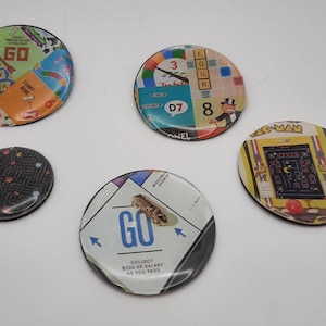 May include: Five round, colorful game board coasters with various designs. The coasters feature images of a game board with the words "GO", "FUNNY MONEY", "3", "FOUR", "D7", "8", "NEL", "PAC-MAN", and "COLLECT $200.00 SALARY AS YOU PASS".