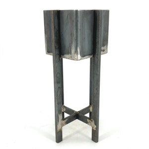 Könnte beinhalten: Ein handgefertigter Metall-Pflanzkübel im Industriestil. Der Pflanzkübel hat ein einzigartiges, vielseitiges Design mit einer dunkelgrauen, rohen Stahloberfläche. Er steht auf vier Beinen, die durch eine X-förmige Stütze verbunden sind, was eine moderne, minimalistische Ästhetik schafft.