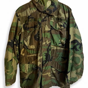 May include: A camouflage field jacket featuring a woodland pattern in shades of green, brown, and black. The jacket includes a high collar, a front zipper closure, and four large button-flap pockets. The text JOHNSON and U.S. ARMY are embroidered.
