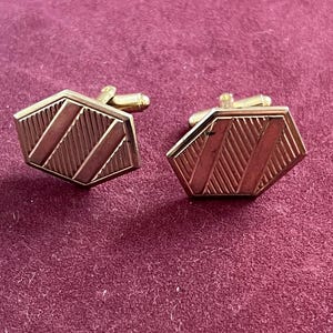 May include: A pair of gold-toned metal cufflinks featuring a hexagonal shape. The surface of each cufflink displays a textured pattern of diagonal stripes alternating with smooth, polished bands. Each piece includes a standard swivel toggle back.