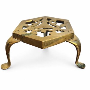 May include: A hexagonal brass trivet featuring an intricate openwork cutout pattern on the top surface. The piece is supported by three curved legs that extend outward from each corner, providing a raised platform with a metallic finish.