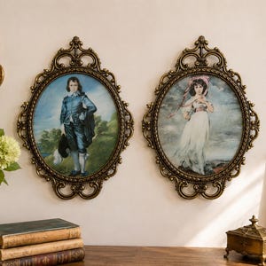 May include: Two oval wall portraits in ornate, antique-style gold frames. The left portrait depicts a person in a blue suit holding a hat, while the right portrait shows a person in a white dress with a pink headpiece, both set against landscapes.