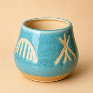 May include: A turquoise ceramic vessel with a rounded shape and a wide opening. The exterior features two abstract, light-colored designs. The base and interior are a warm, light brown. The vessel is likely handmade and suitable for decorative use.