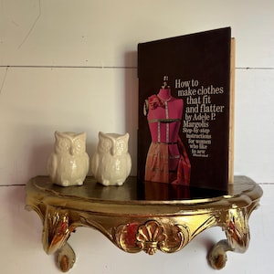 May include: A gold-toned wall shelf with ornate carved details, two white ceramic owl figurines, and a hardcover book titled How to make clothes that fit and flatter by Adele P. Margolis with a pink dress form illustration on the cover.