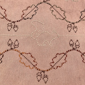 May include: A close-up of a pink textile with embroidered details. The design features a squirrel, oak leaves, and acorns in brown, white, and orange thread. The bottom edge has a fringe.