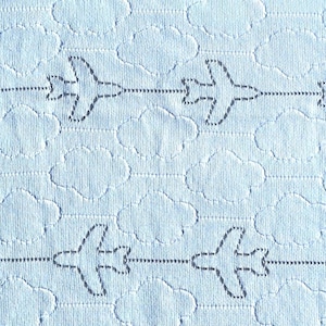 Puede incluir: Tela azul claro con un patrón repetitivo de aviones y nubes. Los aviones están delineados en gris oscuro y conectados por líneas punteadas, creando un diseño temático de viaje. Las nubes también están delineadas.