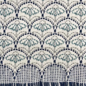 May include: Close-up of a woven textile with a repeating pattern of stylized fan shapes. The design incorporates shades of white, teal, gray, and navy blue. The bottom edge features a fringe in navy blue and white.