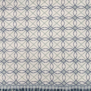 Op de afbeelding: Een wit geweven deken met een geometrisch patroon van blauwe en groene cirkels. De deken heeft een franje rand.