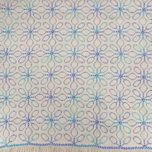May include: A woven textile with a repeating floral pattern in shades of blue and teal on a cream background. The edges are finished with a fringe. The design is a geometric floral pattern.