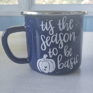 May include: A blue enamel mug with white speckles and a white design that says "tis the season to be basic" with a pumpkin graphic.